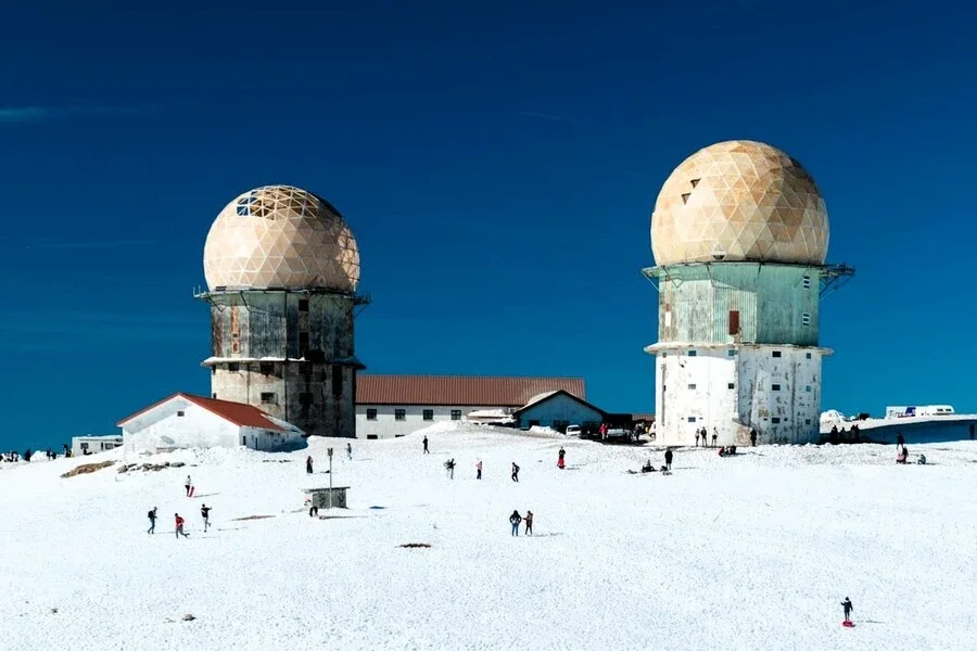 As duas torres de radar icônicas no topo da Serra da Estrela, rodeadas por um manto de neve sob um céu azul profundo, com pessoas caminhando.