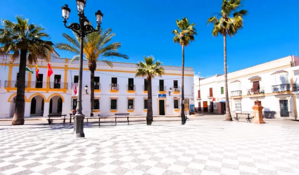 Fachada branca do Ayuntamiento de Trebujena em destaque numa praça com chão axadrezado, palmeiras e céu azul.