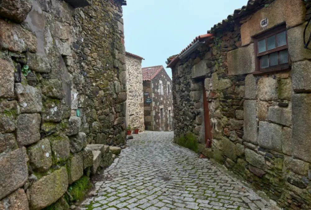 Vista de uma rua estreita e tradicional com casas de granito e pavimento em pedra, típica das aldeias históricas do concelho do Sabugal.
