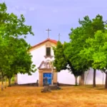 Fachada frontal da Capela de Sacaparte em Alfaiates, um edifício branco com porta azul, emoldurado por árvores verdes e encimado por uma cruz de pedra simples.