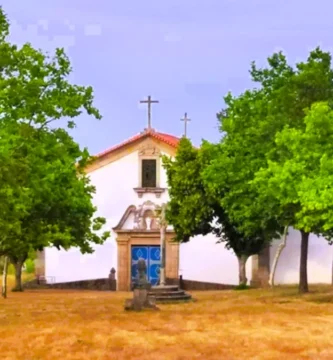 Fachada frontal da Capela de Sacaparte em Alfaiates, um edifício branco com porta azul, emoldurado por árvores verdes e encimado por uma cruz de pedra simples.