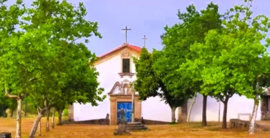 Fachada frontal da Capela de Sacaparte em Alfaiates, um edifício branco com porta azul, emoldurado por árvores verdes e encimado por uma cruz de pedra simples.
