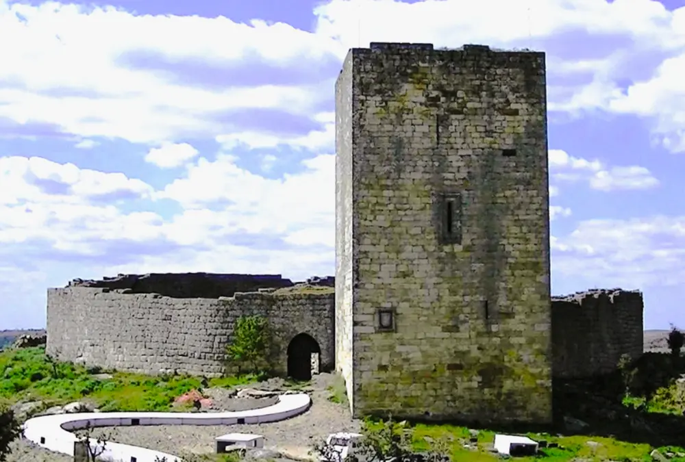 Torre de menagem quadrangular e muralhas circulares do Castelo de Vilar Maior sob céu azul com nuvens.