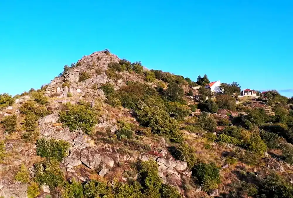 Paisagem serrana verdejante em Vila do Touro, com uma pequena casa de pedra isolada no horizonte sob um céu nublado.
