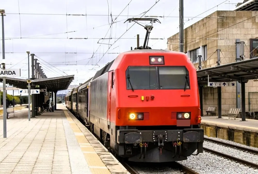 Comboio moderno vermelho parado junto ao cais da estação da Guarda, com sinalética de linhas visível.