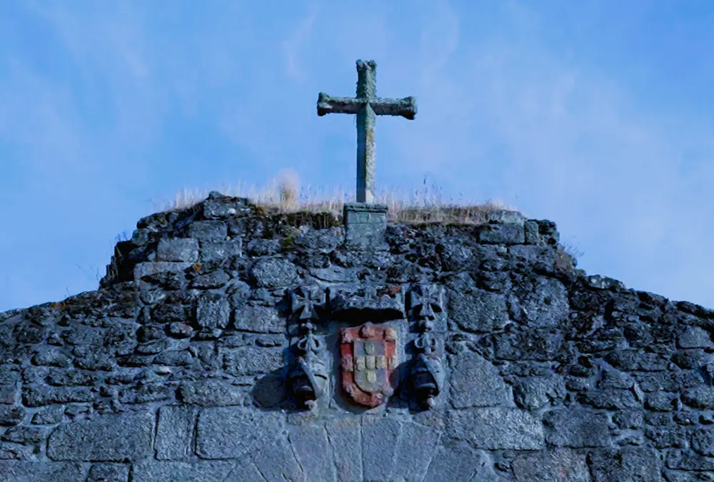 Cruz de pedra no interior do Castelo de Alfaiates, Sabugal, com céu azul intenso ao fundo, marcando o antigo cemitério da vila.
