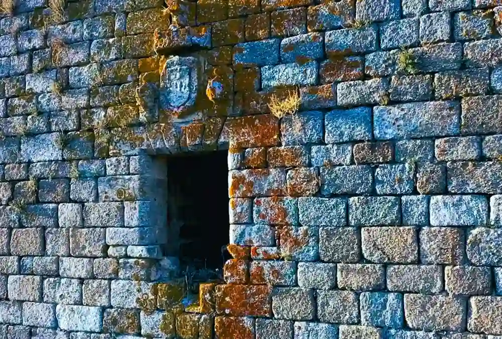 Detalhe de uma janela de granito na muralha do Castelo de Alfaiates com um escudo real esculpido em pedra e líquenes cor de laranja.