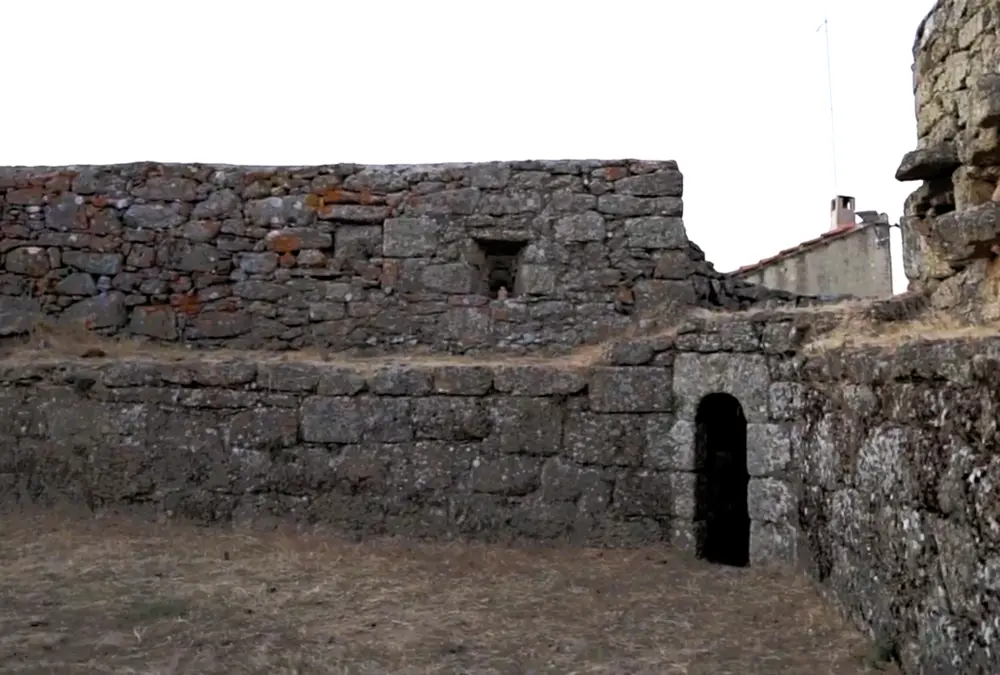 Primeiro plano de um muro de pedra de granito antigo e texturizado com líquenes cor de laranja e amarelos, focado numa pequena abertura quadrada de janela. O termo árabe 'al-ha'it', que significa 'o muro', descreve este tipo de fortificação robusta.