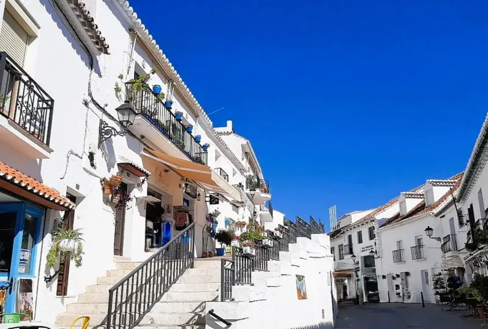 Rua pedonal estreita em Mijas Pueblo com edifícios de arquitetura tradicional andaluza pintados de branco, escadas de pedra e vasos azuis com flores nas varandas sob um céu azul limpo.
