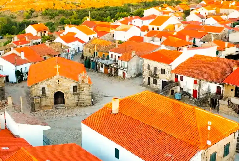 Vista aérea da vila de Alfaiates, no Sabugal, destacando o casario de telhados laranjas e a pequena igreja de pedra na praça central.