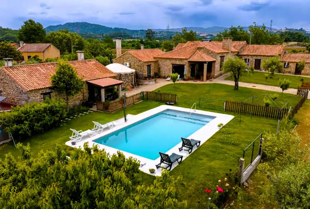 Piscina rodeada por casas de pedra com telhados de telha e áreas verdes, num refúgio rural de Belmonte.
