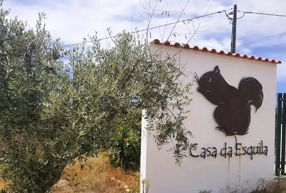 Entrada do restaurante Casa da Esquila no Casteleiro com logótipo de esquilo na parede branca e oliveira ao lado.