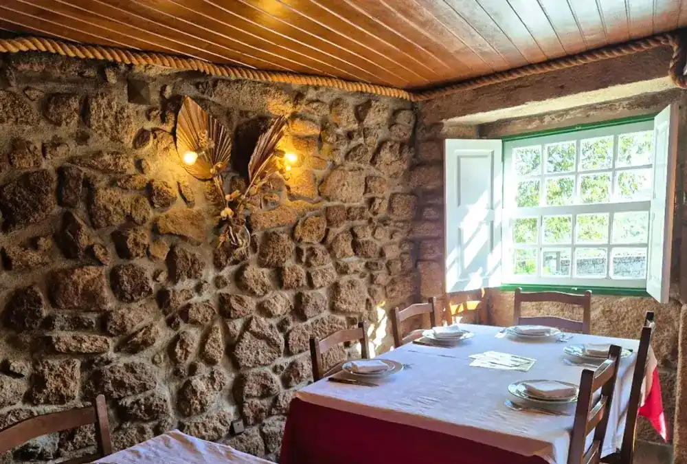 Interior rústico do Restaurante Dom Sancho em Sortelha com paredes de pedra granito, janela de madeira e mesa posta.