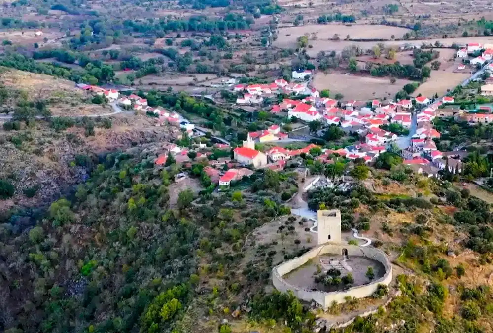 Vista aérea da vila histórica de Vilar Maior, Portugal, mostrando o castelo medieval, a igreja matriz e o casario da aldeia rodeado por paisagem verde e formações rochosas.