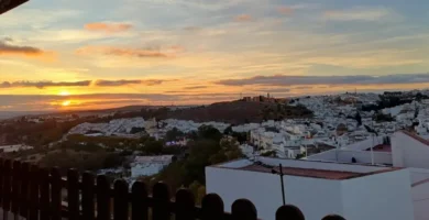 Vista panorâmica de Arcos de la Frontera ao pôr do sol, mostrando o casario branco empilhado na colina sob um céu com tons dourados e azulados.