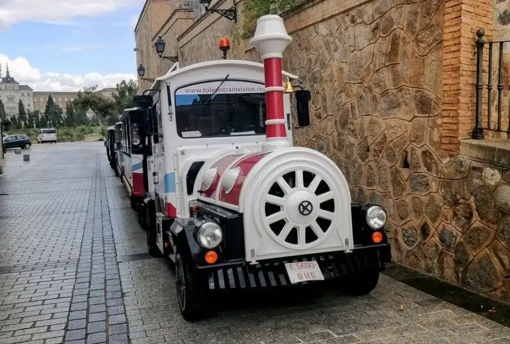 O comboio turístico branco e vermelho de Toledo estacionado numa rua calcetada junto a um muro de pedra histórico, com a inscrição "Toledo Train Vision".