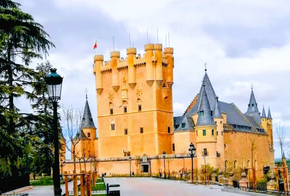 Vista frontal do Alcázar de Segóvia com torre de menagem, telhados de ardósia e bandeira da Espanha, sob céu nublado.