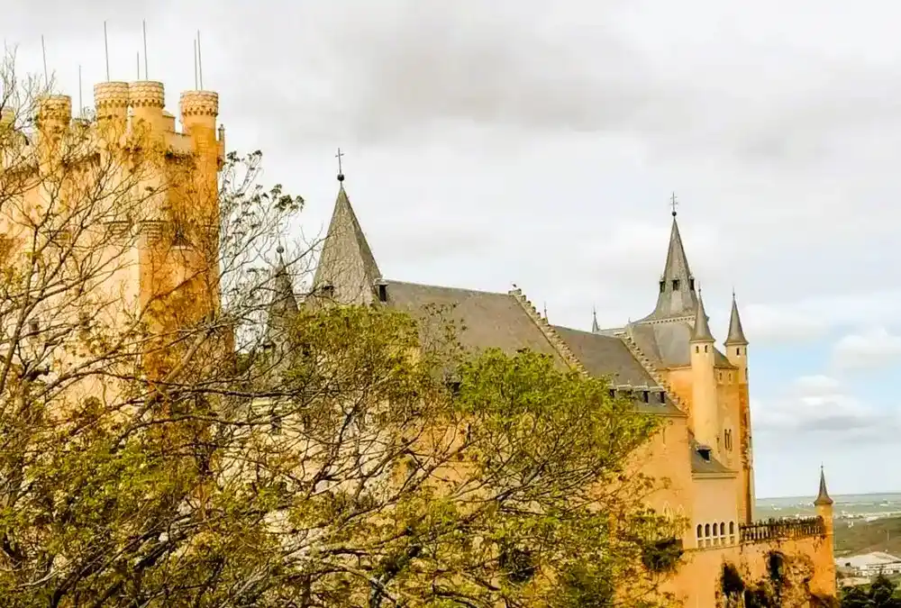 Detalhe das torres cónicas e telhados de ardósia do Alcázar de Segóvia, emoldurado por ramos de árvores. Perspetiva lateral.