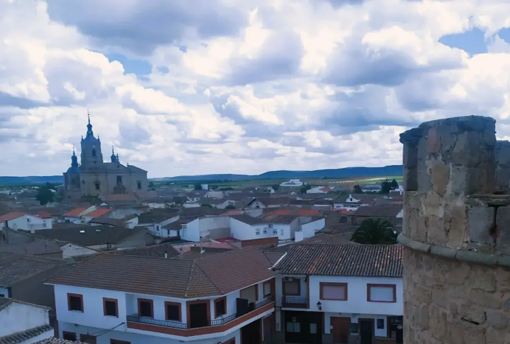 Vista panorâmica sobre os telhados de Almonacid de Toledo, destacando a torre da igreja e a paisagem manchega ao fundo sob um céu nublado.