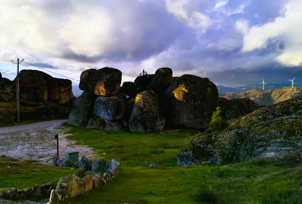 Um grupo de penedos de granito maciços e arredondados em Sortelha, Portugal. A luz dourada do sol da tarde ilumina uma das rochas, contrastando com as nuvens de tempestade dramáticas e as turbinas eólicas visíveis no horizonte.