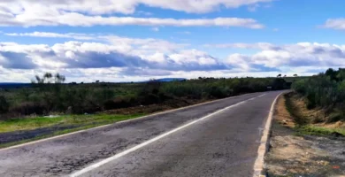 Estrada de asfalto secundária com texturas de desgaste e fissuras estendendo-se em perspectiva central até ao horizonte sob um céu vasto com nuvens cúmulo.