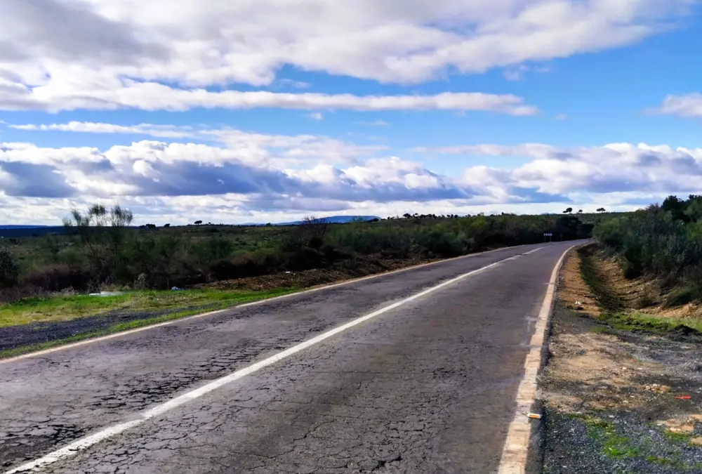 Estrada de asfalto secundária com texturas de desgaste e fissuras estendendo-se em perspectiva central até ao horizonte sob um céu vasto com nuvens cúmulo.