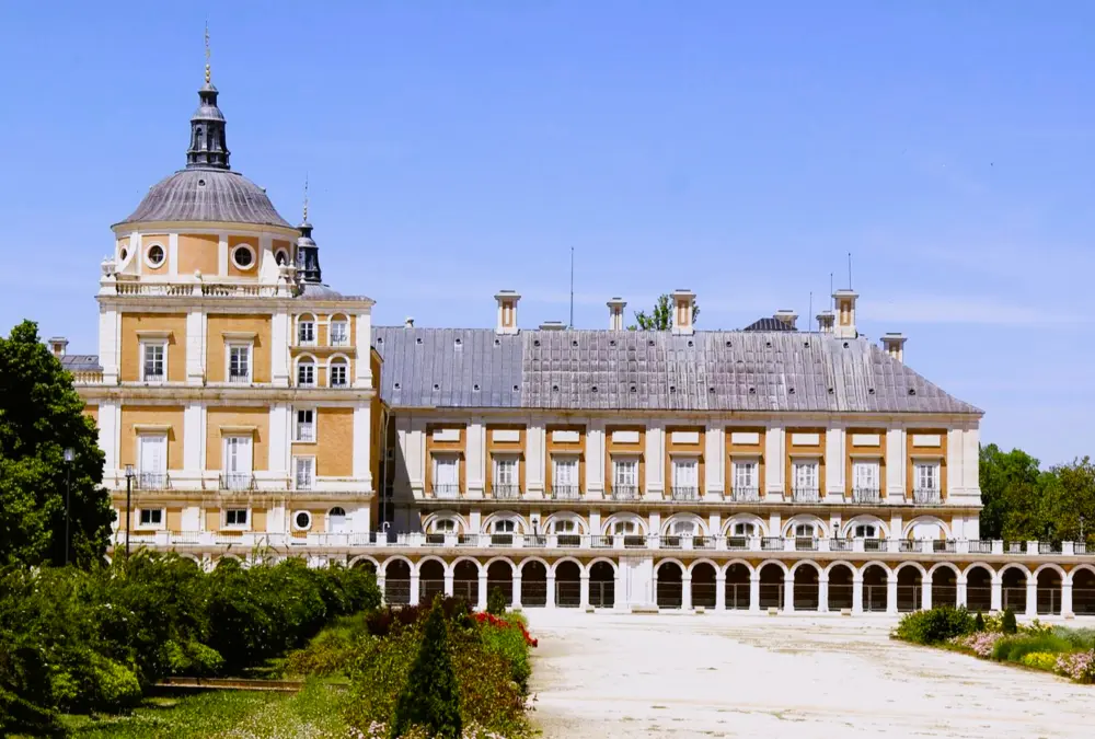 Fachada lateral do Palácio Real de Aranjuez com cúpula e arcadas brancas sob céu limpo.