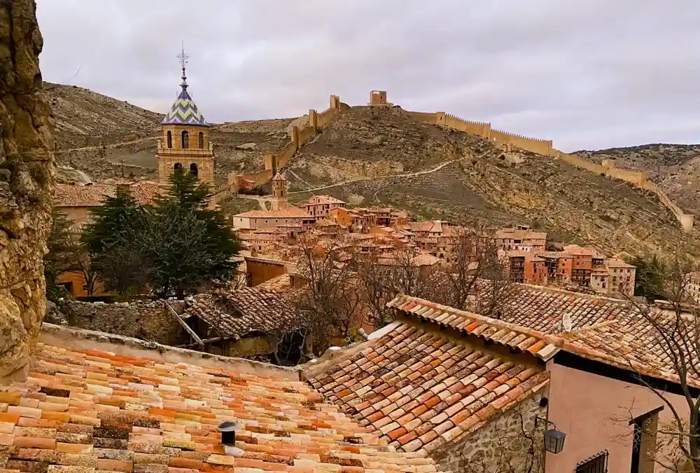 Vista panorâmica dos telhados de Albarracín com a torre da igreja em destaque e a muralha defensiva subindo a colina ao fundo.
