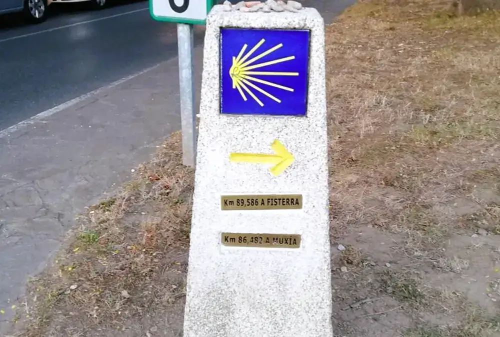 Marco de pedra do Caminho de Santiago com seta amarela e direções para Fisterra e Muxía em estrada secundária.