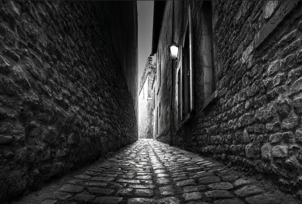 Beco de pedra estreito entre casas antigas, iluminado por uma lamparina na parede, criando sombras fortes e ambiente noturno.
