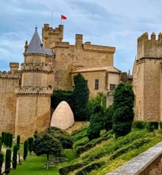 Vista panorâmica das torres e muralhas de pedra clara do Castelo de Olite, com bandeira vermelha no topo, sob um céu nublado e luz natural suave.