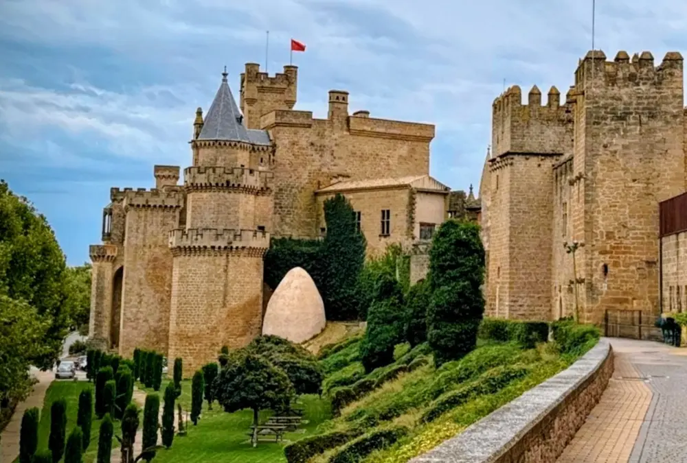 Vista panorâmica das torres e muralhas de pedra clara do Castelo de Olite, com bandeira vermelha no topo, sob um céu nublado e luz natural suave.