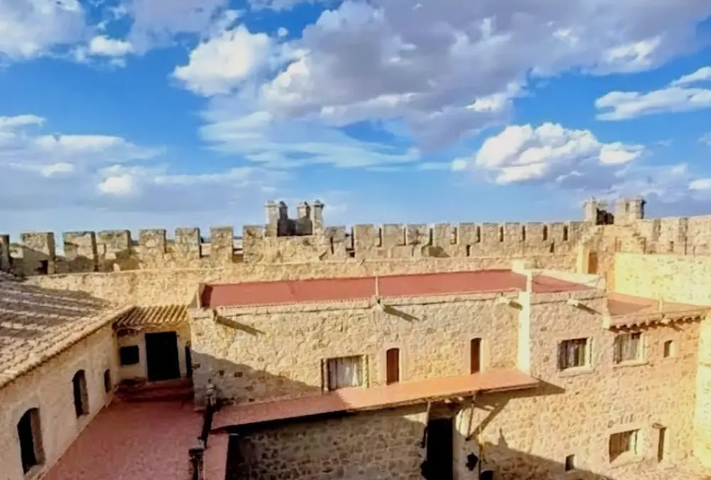 Vista panorâmica do pátio de armas interior do Castelo de Orgaz, Toledo. Estrutura de pedra bem preservada com muralhas merladas, janelas senhoriais e telhados de cerâmica vermelha sob um céu azul com nuvens brancas.