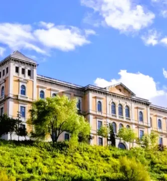 Uma vista panorâmica da colina histórica de Toledo, Espanha, com o edifício da Deputação e as escadas rolantes do Miradero subindo a encosta verde sob um céu azul nublado.