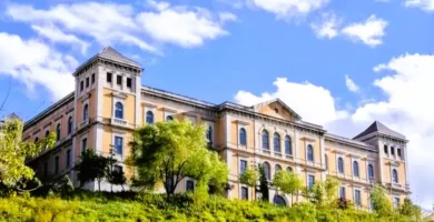 Uma vista panorâmica da colina histórica de Toledo, Espanha, com o edifício da Deputação e as escadas rolantes do Miradero subindo a encosta verde sob um céu azul nublado.