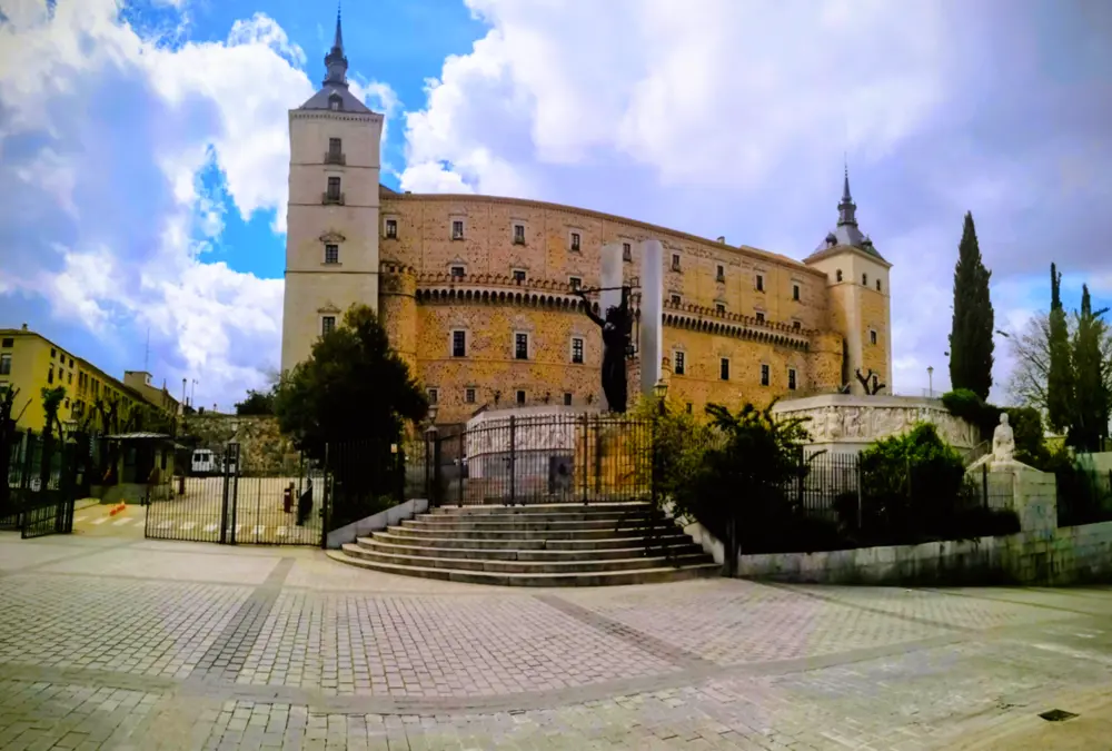 O imponente Alcázar de Toledo, Espanha, um castelo de pedra retangular com quatro torres de canto com pináculos, visto de uma praça calcetada com escadas, uma estátua escura em pé e árvores, sob um céu nublado.