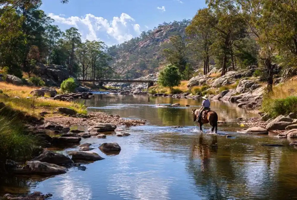 Viajante a cavalo atravessando um rio raso num vale arborizado, exemplo de mobilidade suave e integrada na natureza.