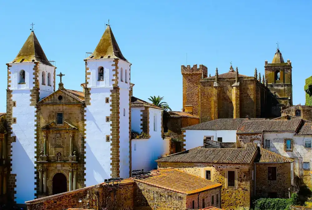Vista panorâmica sobre os telhados de Cáceres, destacando as torres sineiras da Igreja de San Mateo e a Concatedral de Santa María contra um céu azul intenso.