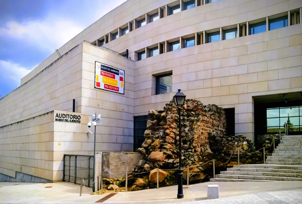 Fachada moderna do Museu do Exército em Toledo, Espanha, integrada com ruínas de pedra antigas e uma escadaria, com um cartaz que diz "Tu Museo, Tu Ejército, Tu Historia".