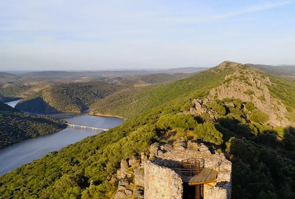 Vista aérea do topo de uma torre de pedra no Parque de Monfragüe, com o rio Tejo e uma ponte ao fundo entre colinas verdes.
