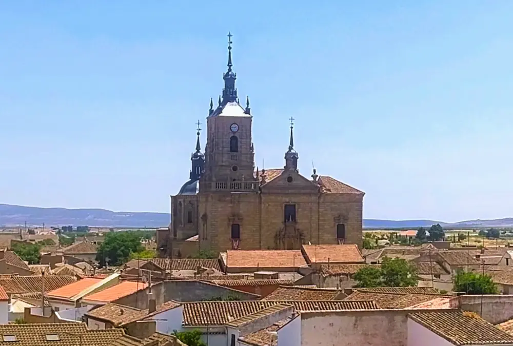 Vista elevada da Igreja de Santo Tomás Apóstol em Orgaz, Toledo. A imponente estrutura de pedra em estilo barroco castelhano ergue-se acima dos telhados de cerâmica da vila, com as suas torres sineiras e cúpulas de metal contra um céu azul claro. Paisagem da Mancha ao fundo.