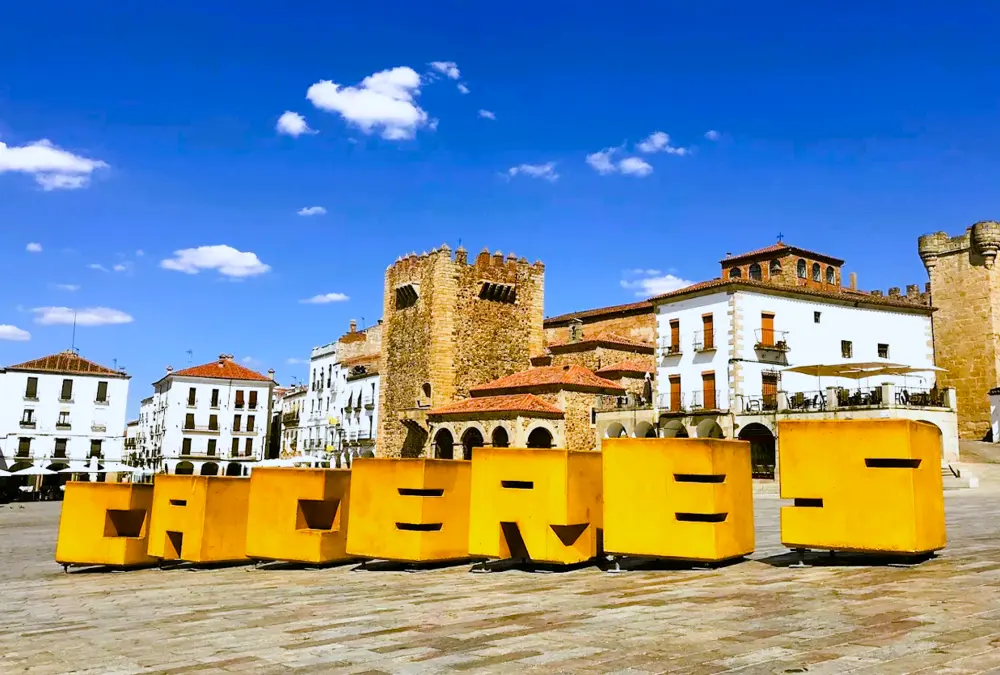 Vista vibrante da Plaza Mayor de Cáceres, com a Torre de Bujaco e as arcadas medievais ao fundo, e o letreiro escultural amarelo "CACERES".