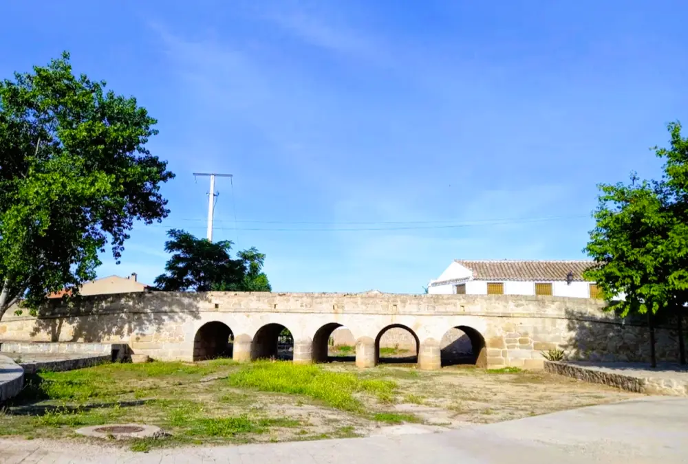 Fotografia da Puente de los Cinco Ojos em Orgaz, Toledo, uma ponte de pedra do século XVIII com cinco arcos de volta inteira. A ponte atravessa um ribeiro seco com vegetação verde, ladeada por árvores e uma casa tradicional branca ao fundo, sob um céu azul.