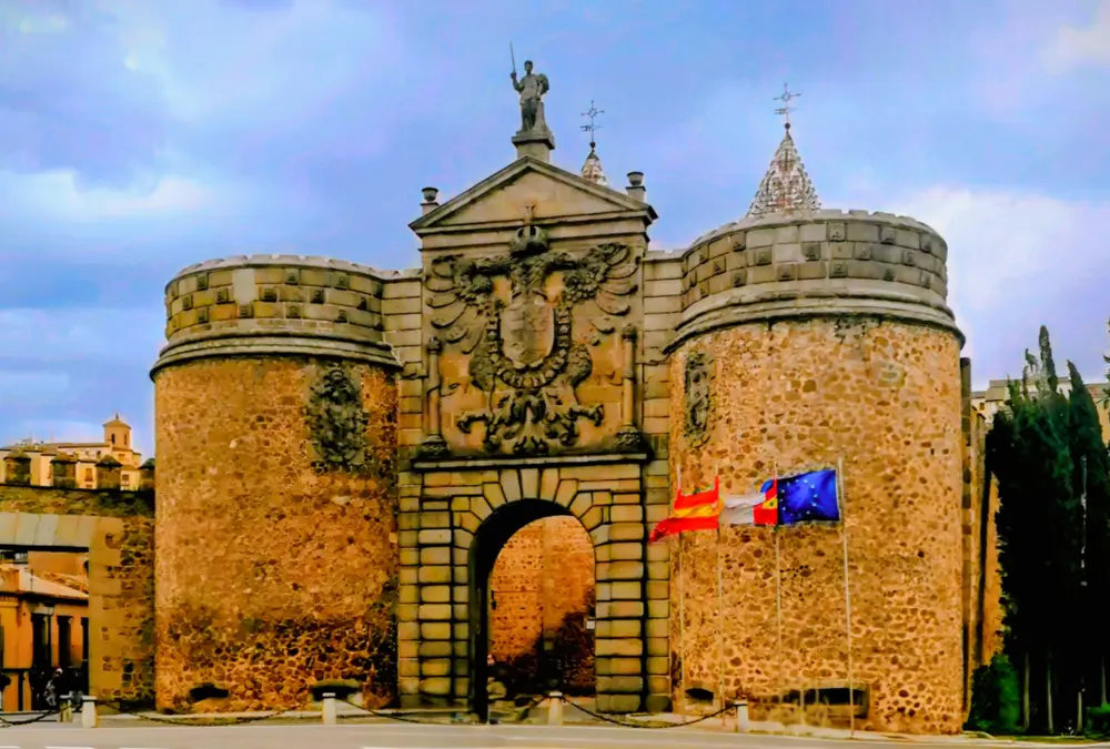 A majestosa Puerta de Bisagra Nueva, em Toledo, Espanha, uma porta monumental de pedra com um arco central, brasão imperial e duas torres redondas de pedra, com bandeiras de Espanha e da UE à frente, sob um céu nublado.
