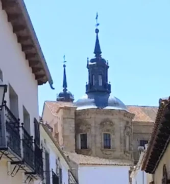 Vista de uma rua estreita e tradicional no centro histórico de Orgaz, Toledo. Fachadas caiadas de branco com varandas de ferro forjado e candeeiros de rua clássicos. Ao fundo, no final da rua, as cúpulas ornamentadas e as torres sineiras da Igreja de Santo Tomás sob um céu azul.
