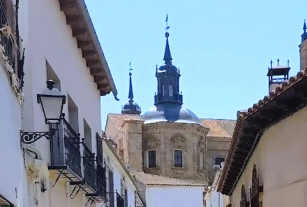 Vista de uma rua estreita e tradicional no centro histórico de Orgaz, Toledo. Fachadas caiadas de branco com varandas de ferro forjado e candeeiros de rua clássicos. Ao fundo, no final da rua, as cúpulas ornamentadas e as torres sineiras da Igreja de Santo Tomás sob um céu azul.