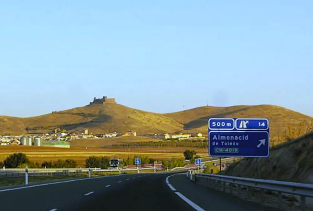 Placa de sinalização na estrada indicando a saída para Almonacid de Toledo com o castelo ao fundo.