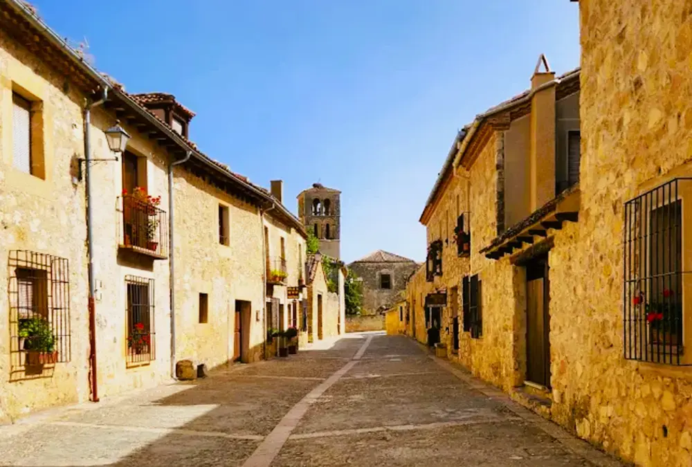 Perspetiva da Calle Real de Pedraza, uma rua empedrada ladeada por edifícios históricos de pedra que conduz à torre da igreja.
