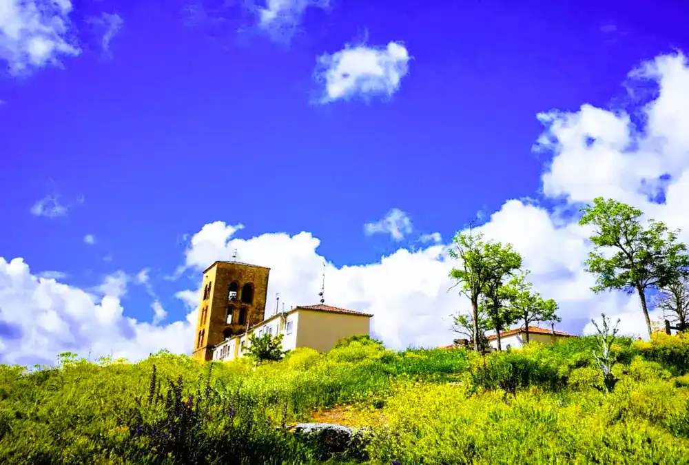 Vista distante e ascendente da torre sineira românica da Igreja de San Salvador em Sepúlveda, que se ergue sobre uma colina verdejante coberta de arbustos e flores silvestres sob um céu azul vibrante com nuvens brancas.