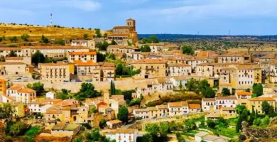 Uma vista panorâmica aérea da vila medieval de Sepúlveda, em Castela e Leão, Espanha, aninhada numa colina de pedra dourada com o desfiladeiro das Hoces del Duratón e campos rurais que se estendem ao fundo sob um céu nublado.